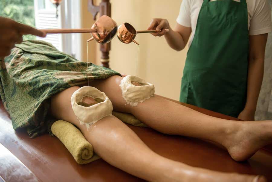 A patient receiving Janu Vasti, a traditional Ayurvedic treatment for joint pain, with practitioners pouring warm medicated oil from copper ladles into dough rings placed over both knees.